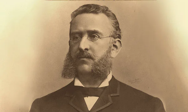 Photographie vintage avec des lunettes rondes et des favoris distinctifs, portant un costume sombre et un nœud papillon.