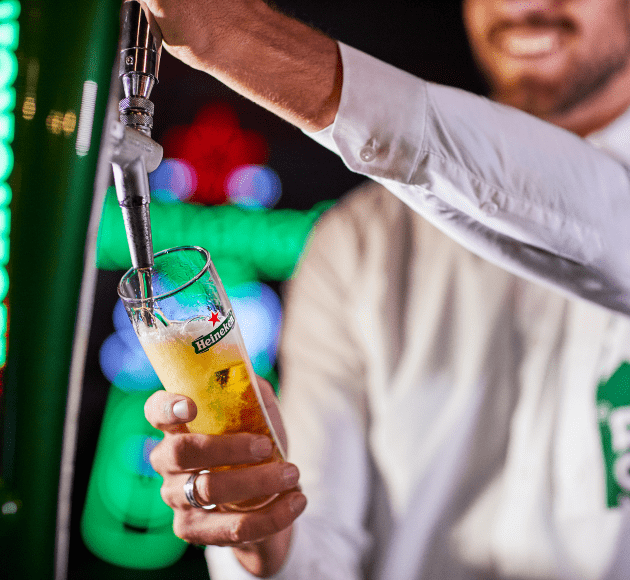 contact_pro Un serveur tire une bière Heineken à la pression dans un verre Heineken, avec un arrière-plan flou de lumières colorées.